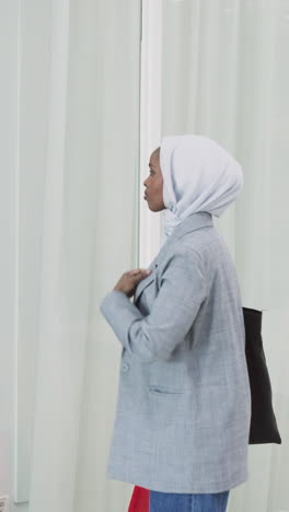 african-american woman adjusts hijab looking at reflection in glass wall slow motion. muslim lady in jacket visits business center. arabic tradition