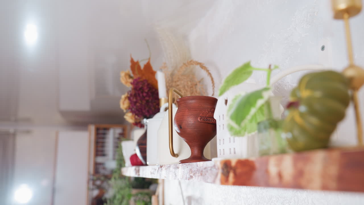 Close-up of neatly arranged flower vessel with dried floral bouquet, wooden goblets, white decorative house, ceramic watering cans, and candle stand on rustic wooden shelf in bright kitchen interior