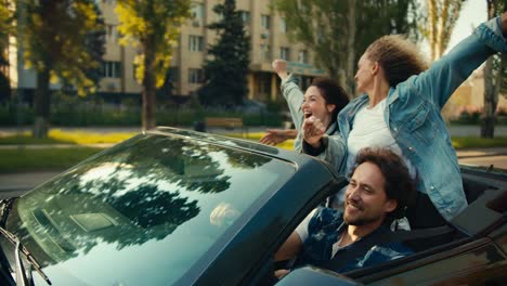 A-happy-trio:-a-guy-and-two-girls-are-riding-in-a-gray-convertible.-A-blonde-girl-and-a-brunette-girl-have-fun-and-rejoice.-Happy-cabriolet-ride