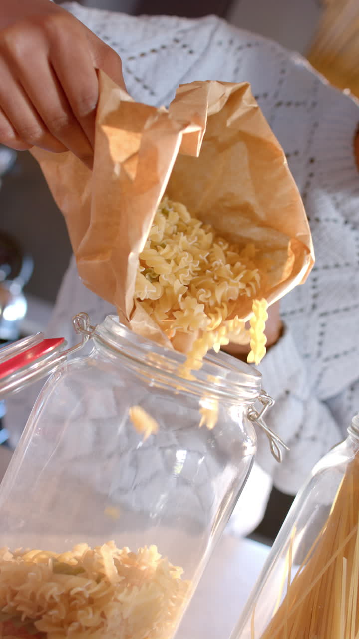Vertical video of midsection of african american woman pouring pasta to jar in kitchen, slow motion