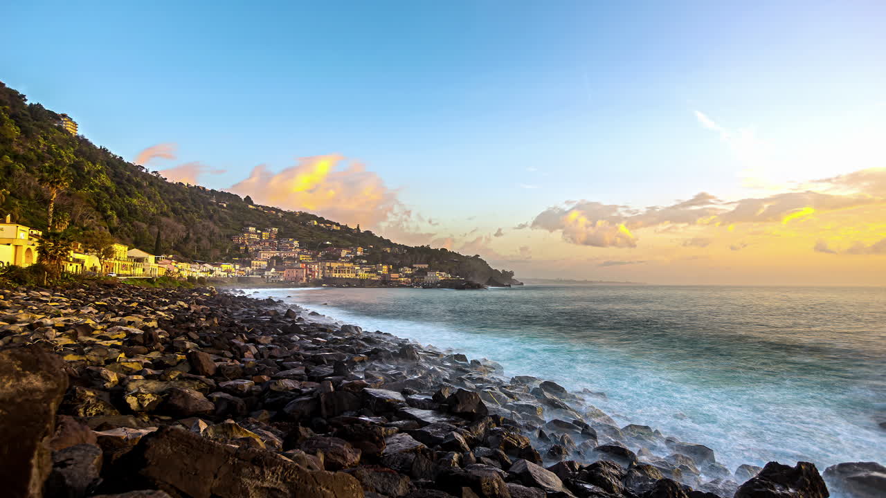 foto panoramica della città di acireale in sicilia con onde che raggiungono la costa rocciosa e raggi dorati del sole che illuminano gli edifici situati sul pendio della montagna