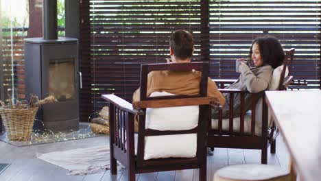 Mixed-race-couple-toasting-and-drinking-coffee-while-sitting-near-the-fireplace-at-vacation-home