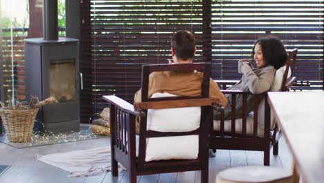 pareja de raza mixta brindando y bebiendo café mientras están sentados cerca de la chimenea en la casa de vacaciones