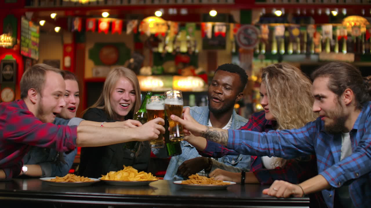 In the company of funny dresies the girl says a toast and everyone checks their glasses with beer and smiles while sitting in a bar with men and women. Party of modern students..