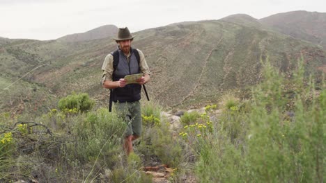 Un-Hombre-Caucásico-Barbudo-Que-Lee-Un-Mapa-De-Supervivencia,-Caminando-Por-Un-Sendero-De-Montaña-En-El-Desierto
