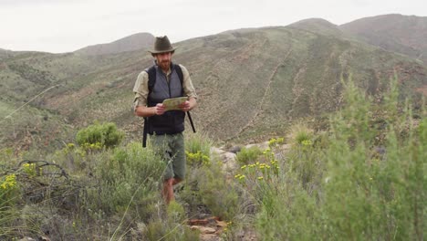 hombre caucásico barbudo sobreviviente leyendo un mapa, caminando por un sendero de montaña en el desierto