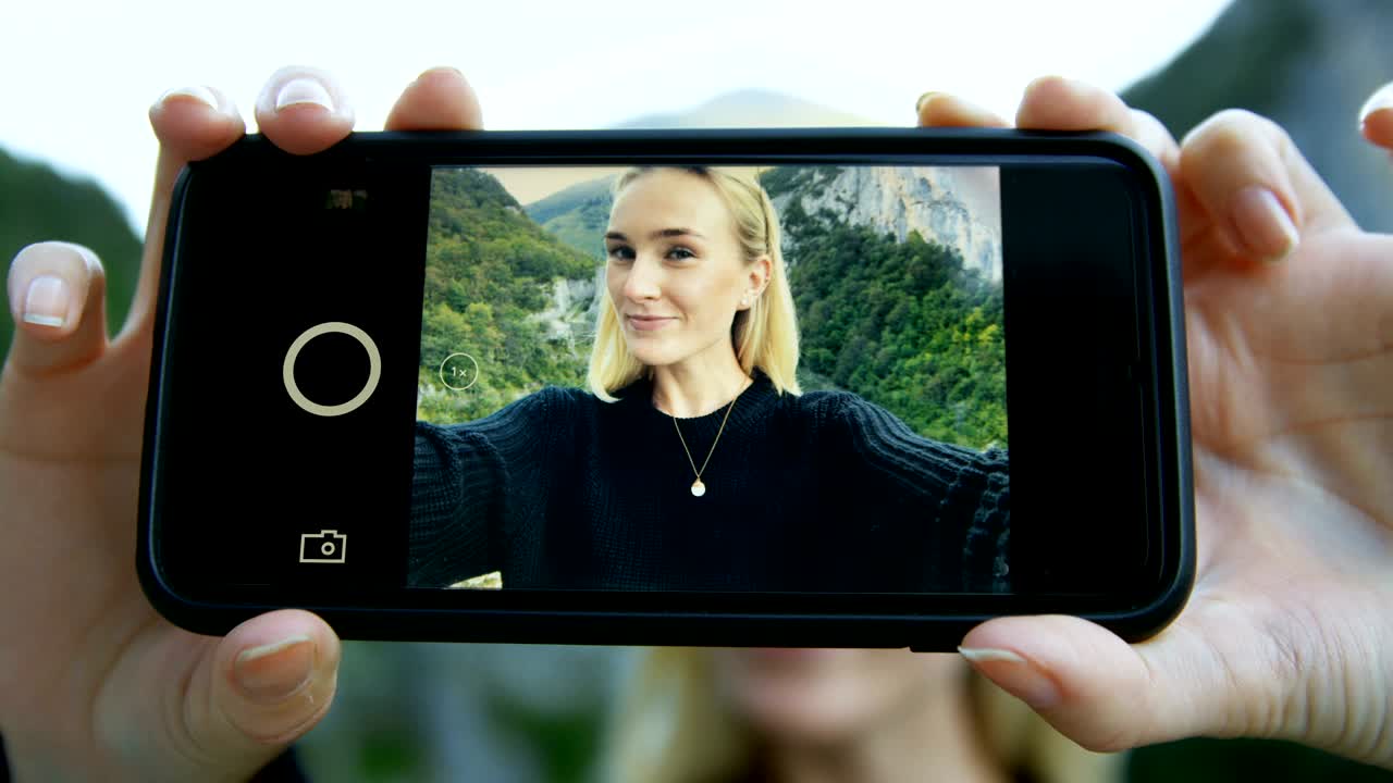 primer plano en la pantalla del teléfono inteligente de la hermosa chica rubia tomando una selfie. en el fondo pintorescas colinas verdes y vistas a las montañas.