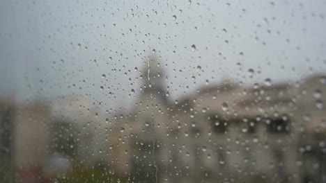 Una-Vista-En-Cámara-Lenta-De-Las-Gotas-De-Lluvia-Vistas-A-Través-De-Una-Ventana-Durante-Fuertes-Lluvias,-Con-Un-Paisaje-Urbano-Al-Fondo