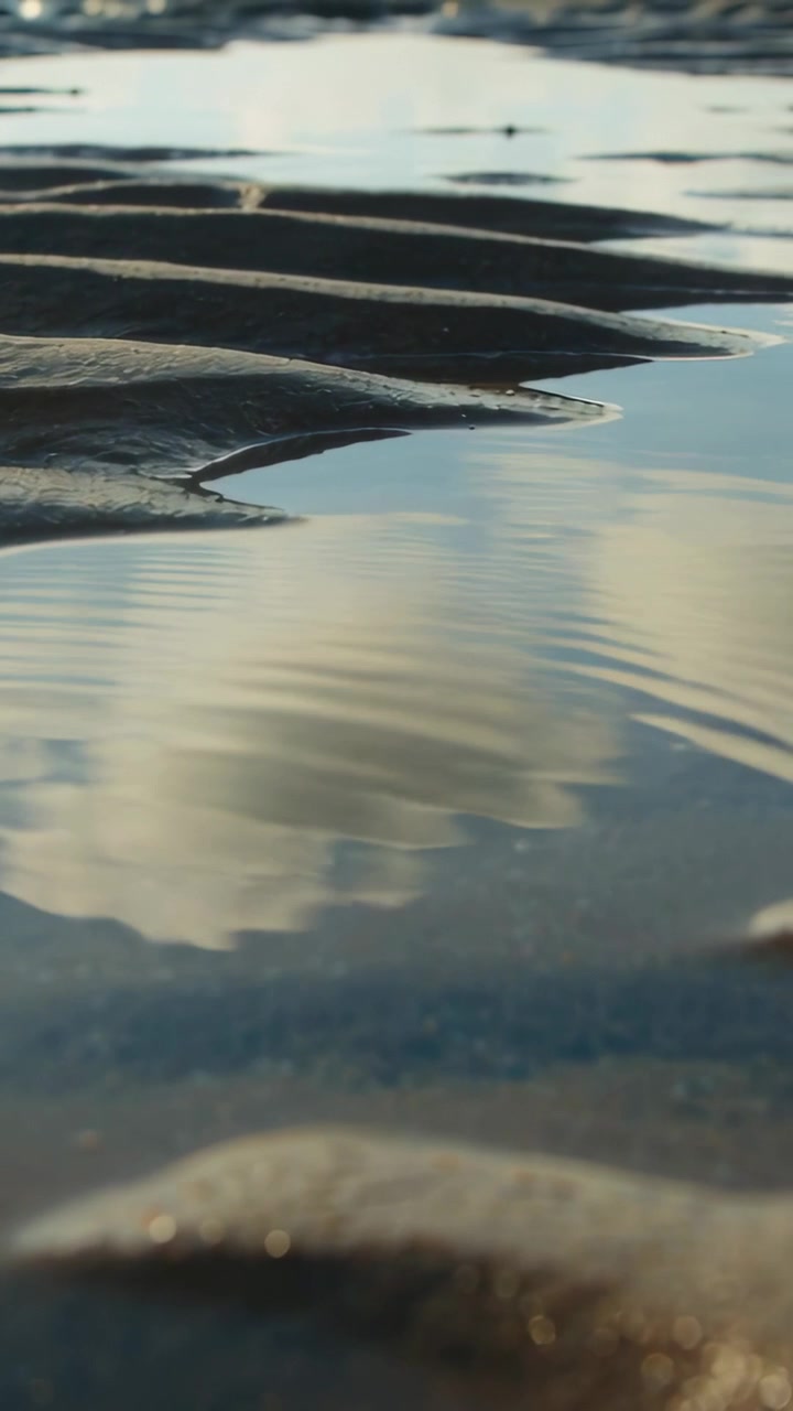 Vertical video: Rippling tidal pool and sand ripples from gentle breeze, reflecting clouds on shore