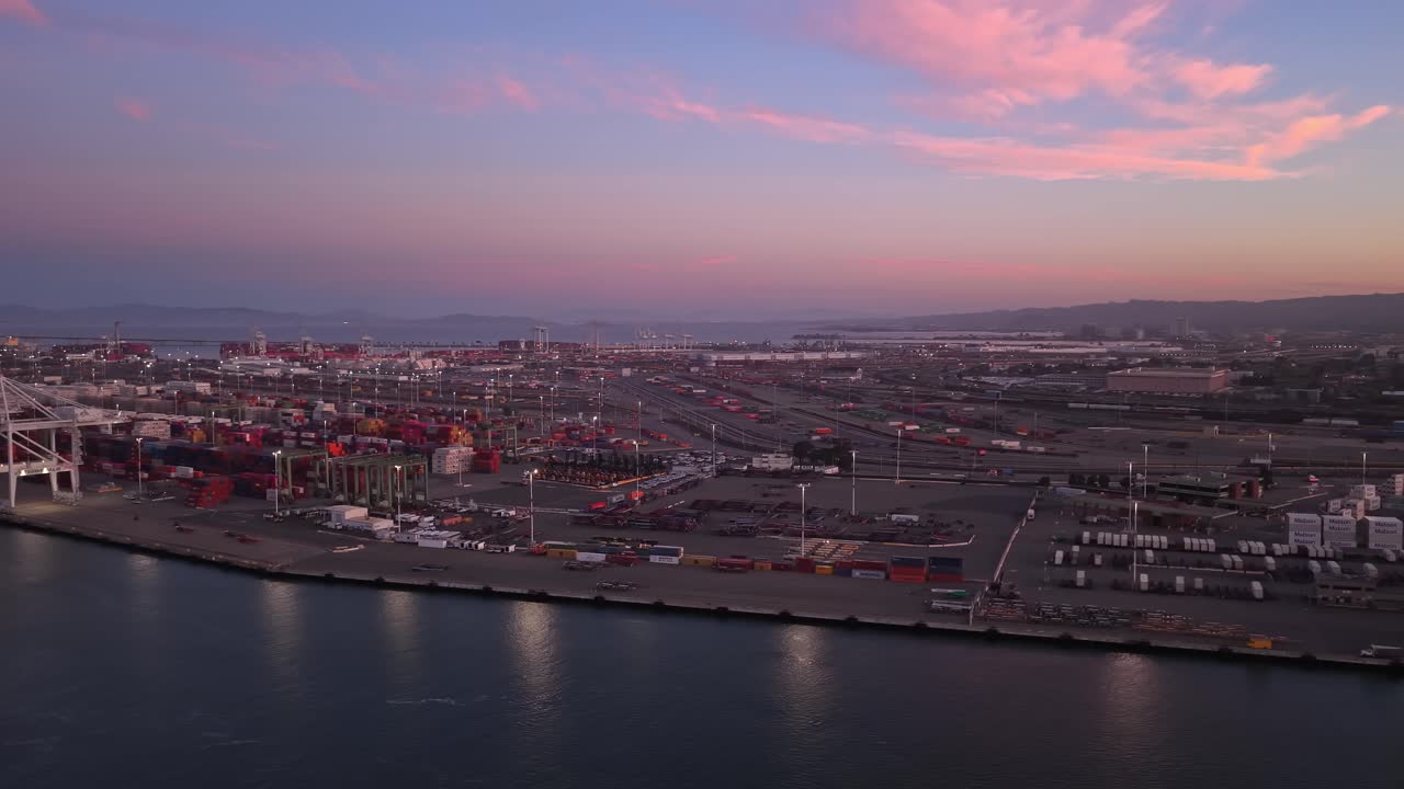 Beautiful aerial view panning across the Port of Oakland in California, showing shipping containers and cranes against a colorful sky with pink clouds during an early morning sunrise
