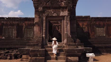 una hermosa mujer con un largo vestido blanco y un sombrero vietnamita subiendo las escaleras al palacio en wat phou arruinó el complejo del templo hindú khmer. champassak, laos, asia. cultura antigua arquitectura religiosa lenta.