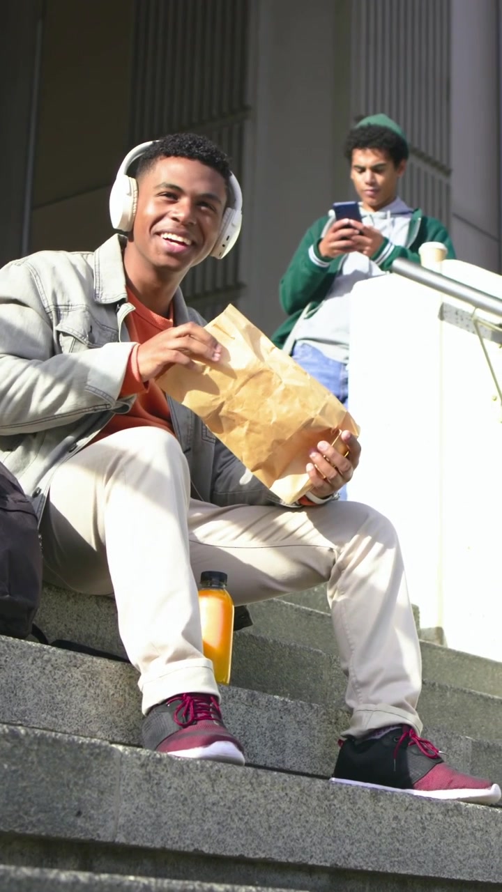 Vertical video: Opening paper bag, man with headset eating, packing up pack on building steps