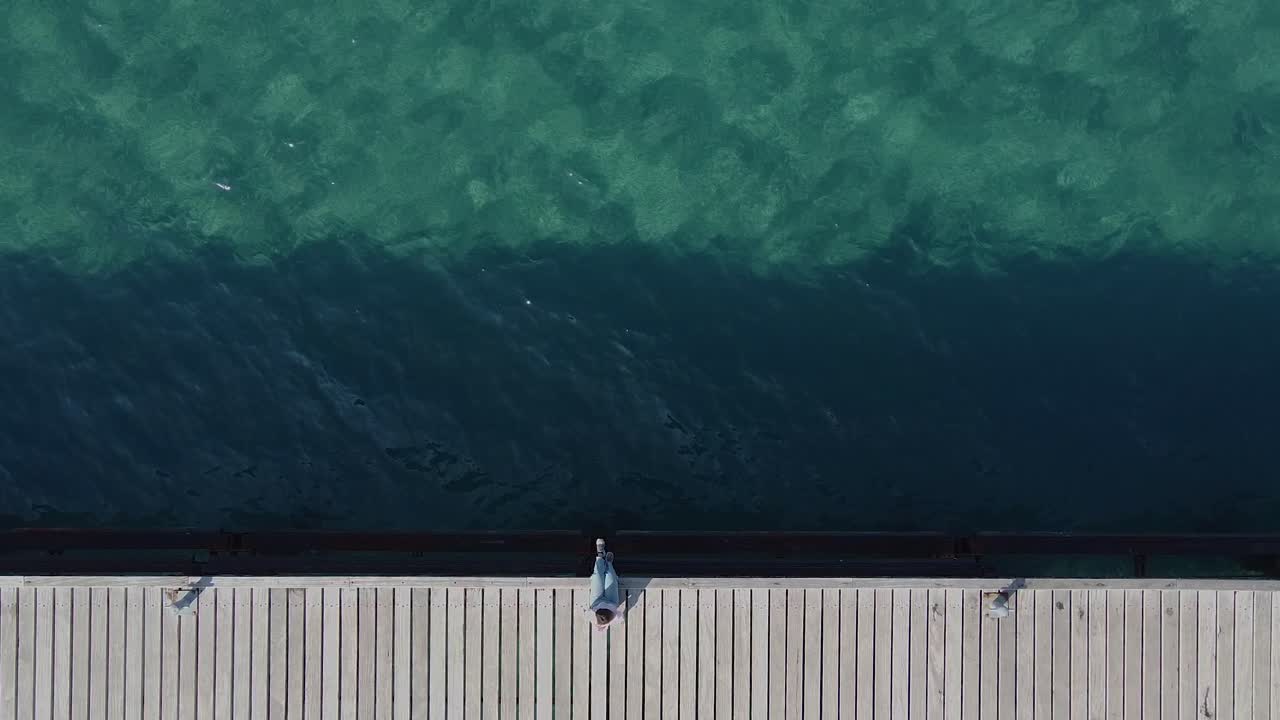 Bird's eye view of a young girl relaxing on the edge of a wooden pier on tropical waters