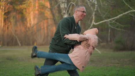 a man in a green jacket and a little girl in a pink jacket, both wearing blue jeans, are joyfully playing together outdoors on a lush green grassy field