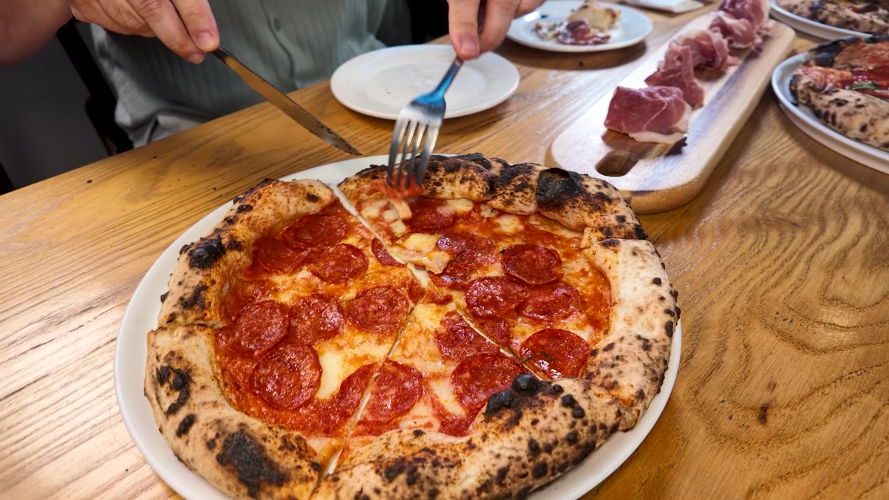 Primo piano di mani che tagliano una pizza al salamino piccante cotta nel forno a legna su un tavolo di legno con salumi.
