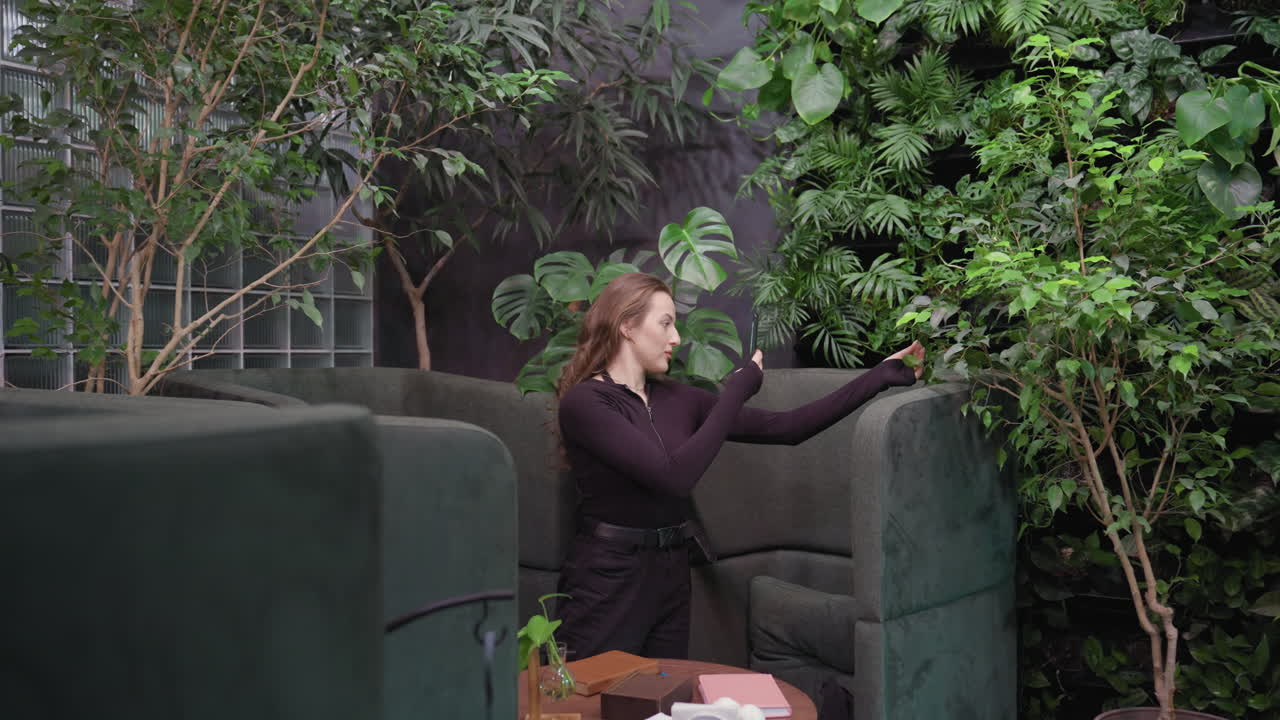 Pretty woman holding phone and adjusting position to photograph lush green leaves while standing near cushioned seating in indoor space decorated with plants, brick wall, and glass backdrop