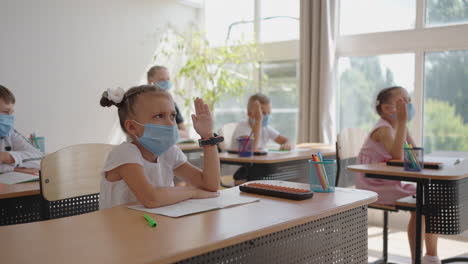 Kinder-Im-Klassenzimmer-Der-Schule-Sitzen-In-Masken-Im-Klassenzimmer-Und-Beantworten-Die-Fragen-Des-Lehrers,-Indem-Sie-In-Zeitlupe-Die-Hände-Heben.-Unterricht-Während-Der-Pandemie-In-Der-Schule