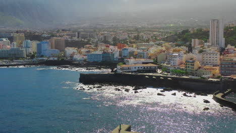 The-vibrant-city-of-Puerto-de-la-Cruz,-Tenerife,-Canary-Islands,-and-its-harbor-with-stone-piers,-rocks-and-reefs-washed-by-the-waves-of-the-Atlantic-ocean-on-a-sunny-summer-day,-zoom-aerial-shot-4K