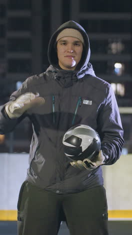 hombre con capucha y guantes sosteniendo una pelota de fútbol pensativamente bajo las luces nocturnas en el campo deportivo urbano, edificios de gran altura y poste de la portería en el fondo