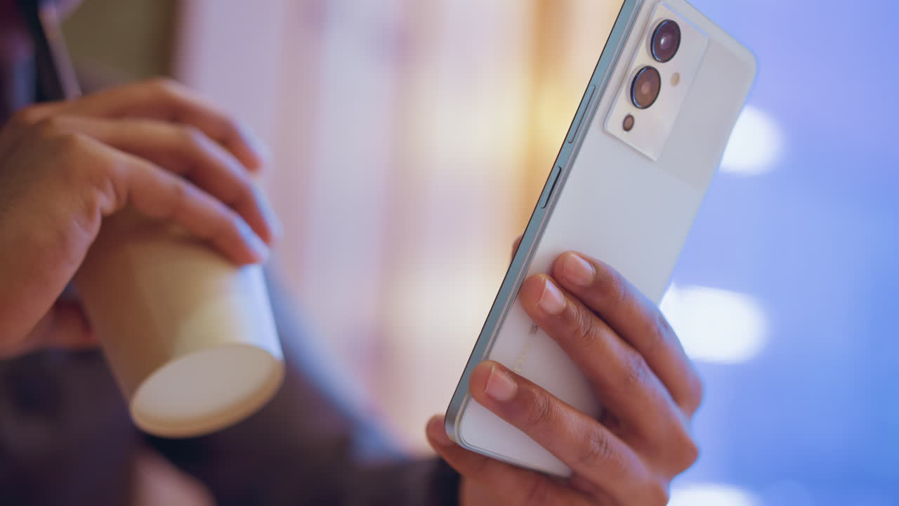 Close-up of hand holding smartphone near beverage cup in softly lit environment with blurred bokeh lights in background, suggesting relaxed atmosphere, digital interaction, and modern casual lifestyle indoors