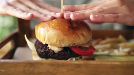 Tasty-burger-with-french-fries-on-a-wooden-tray.-Female-hands-touching-it-to-start-taking.-Slow-Motion-shot