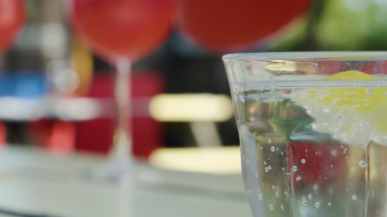 Glass of sparkling water with lemon beside red cocktail, shallow depth, outdoor daylight, camera pans