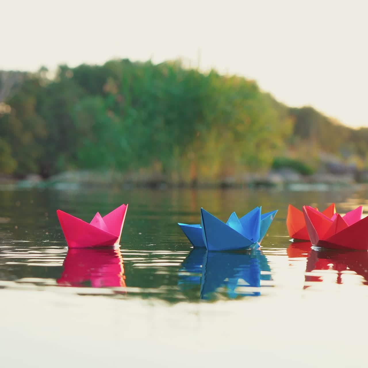 several multi-colored origami in the shape of ships are floating on the water in the lake in the evening on the background of the summer landscape