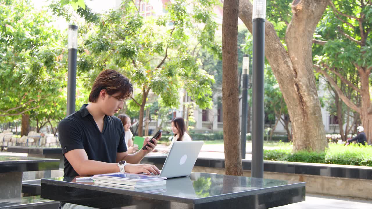 Seorang mahasiswa laki-laki menggunakan laptop dan smartphone sambil duduk di meja kampus luar ruangan