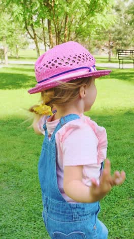 young girl wearing pink hat and denim overalls playing joyfully in a sunny green park setting