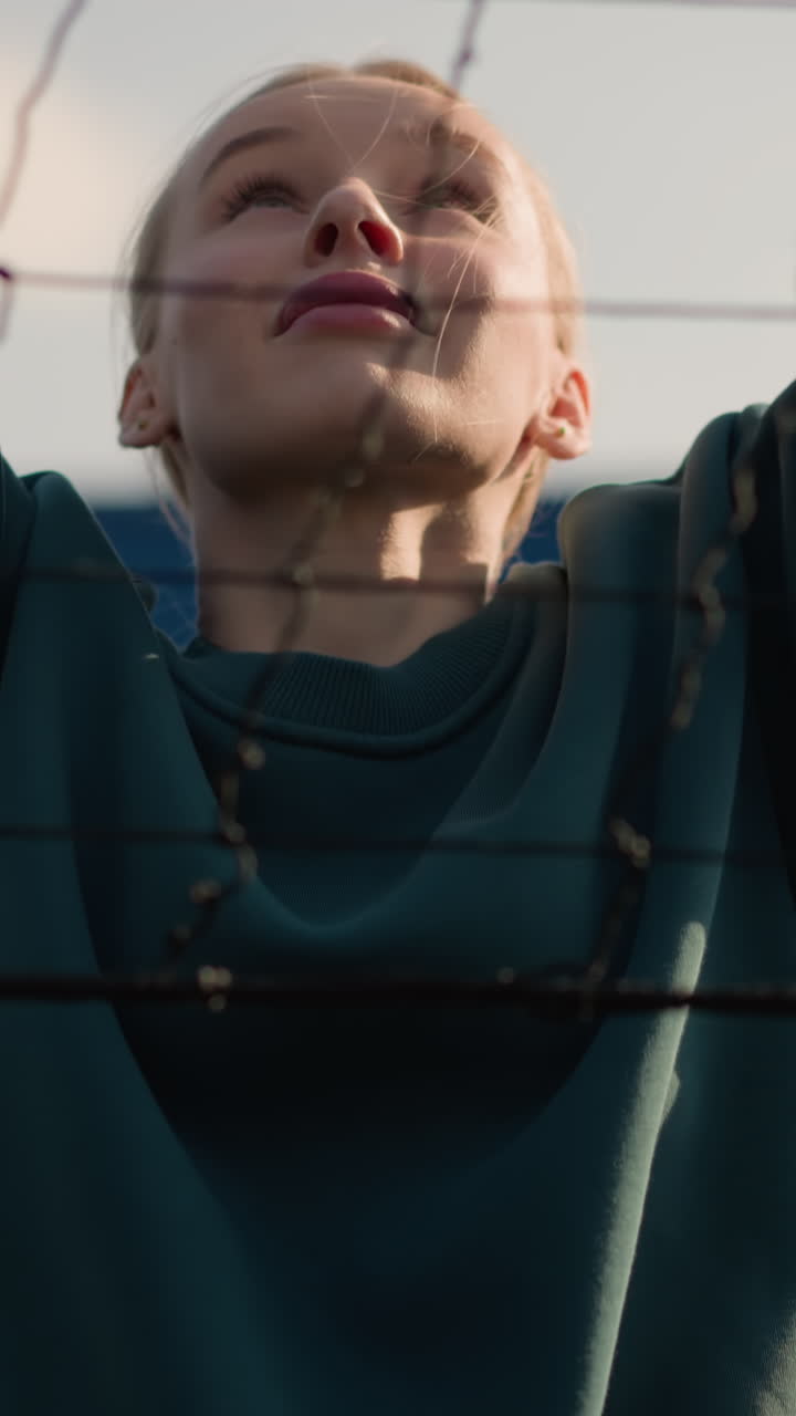 primer plano de una mujer con capucha verde haciendo ejercicio al aire libre, saltando con las manos levantadas cerca de la red, el fondo presenta un edificio borroso y un aro de baloncesto