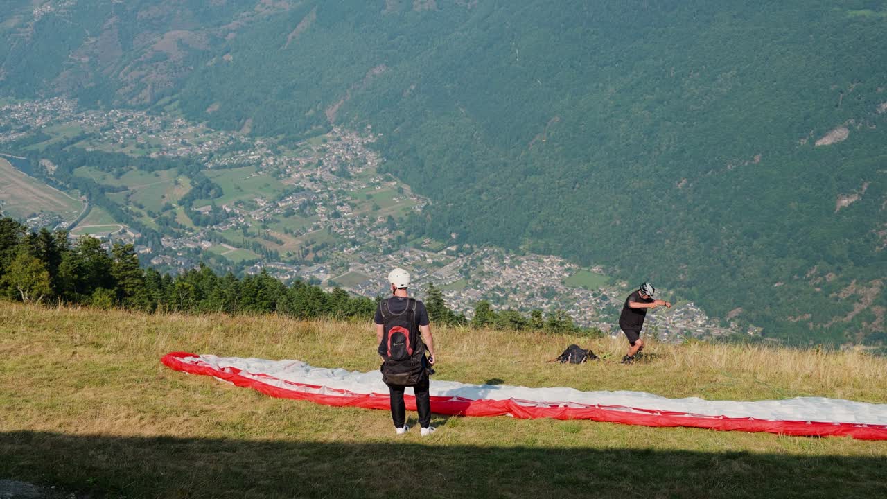 Two paragliders prepare for takeoff on a grassy hillside with a scenic valley view below