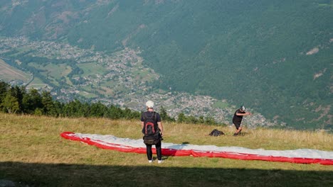 two paragliders prepare for takeoff on a grassy hillside with a scenic valley view below