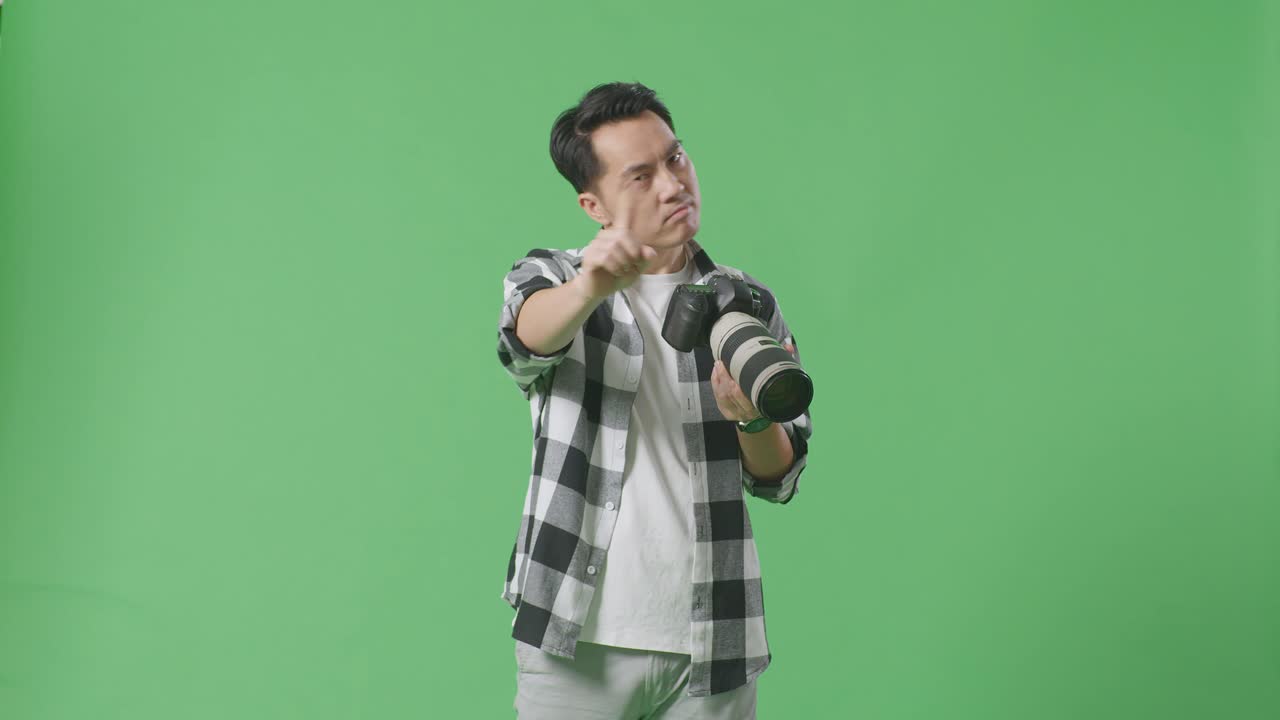 심각한 아시아 사진작가는 손에 카메라를 들고 스튜디오에서 녹색 스크린 배경에 서 있는 동안 지표손가락 표시 없이 불쾌감을 표합니다.
