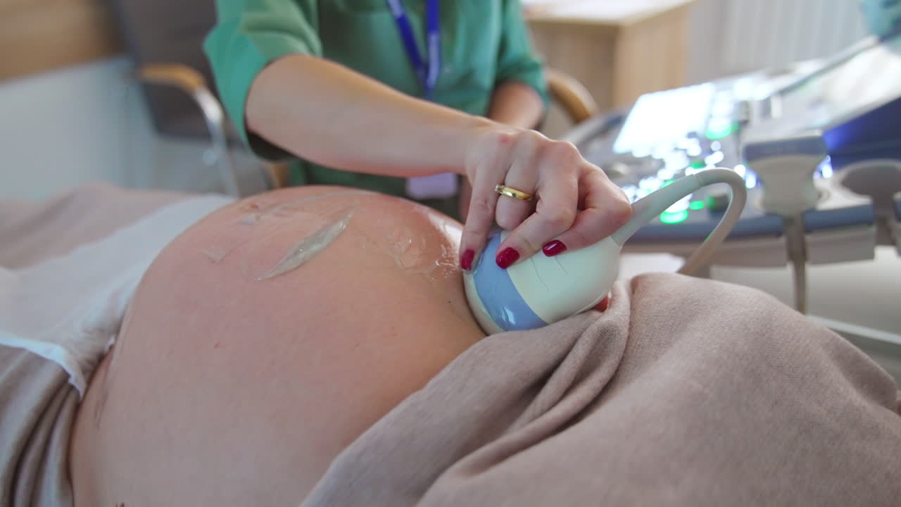 Device is moved by the big pregnant belly for ultrasound diagnostics. Doctor sitting at apparatus at backdrop in blur.