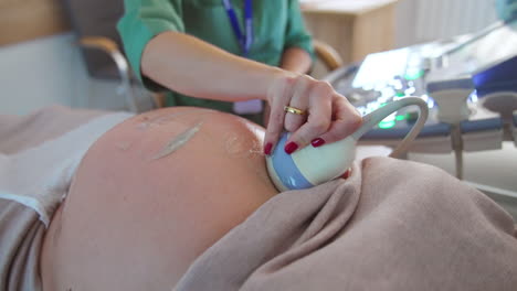 device is moved by the big pregnant belly for ultrasound diagnostics. doctor sitting at apparatus at backdrop in blur.