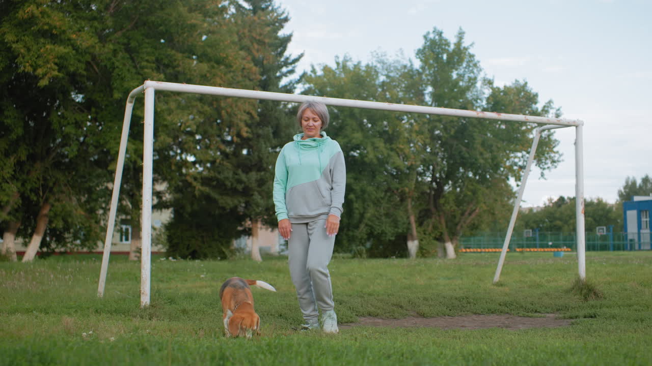 Dog owner performing knee bend exercise near goal post while dog moves around on grassy field during outdoor workout session showcasing fitness, pet companionship, and active healthy lifestyle