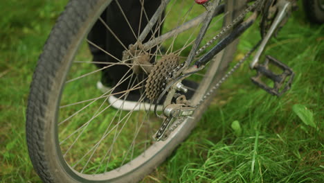 close-up of someone standing next to parked bicycle in grassy field, adjusting the pedal backward, lifting the back tire, pedaling to rotate the wheels, and gently lowering the bike back down