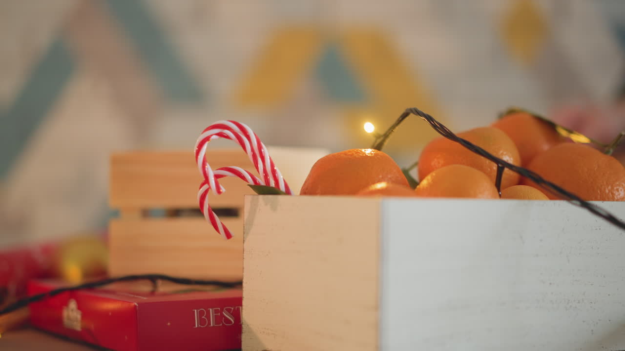 Crate oranges candy canes christmas lights, wooden box filled tangerines and striped candy canes, warm fairy lights creating soft bokeh, red wrapping paper peeking underneath, cozy rustic holiday