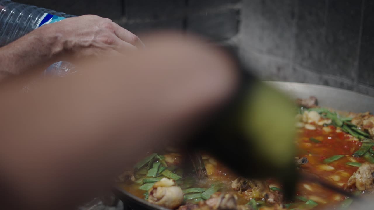 Preparing Paella Outdoors: Pouring Water into a Large Pan with Chicken and Vegetables
