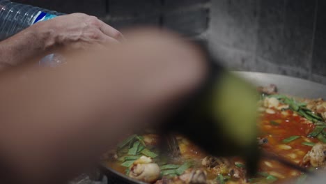 preparing paella outdoors: pouring water into a large pan with chicken and vegetables