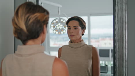 middle-aged woman looking mirror standing in modern cosmetology clinic close up.
