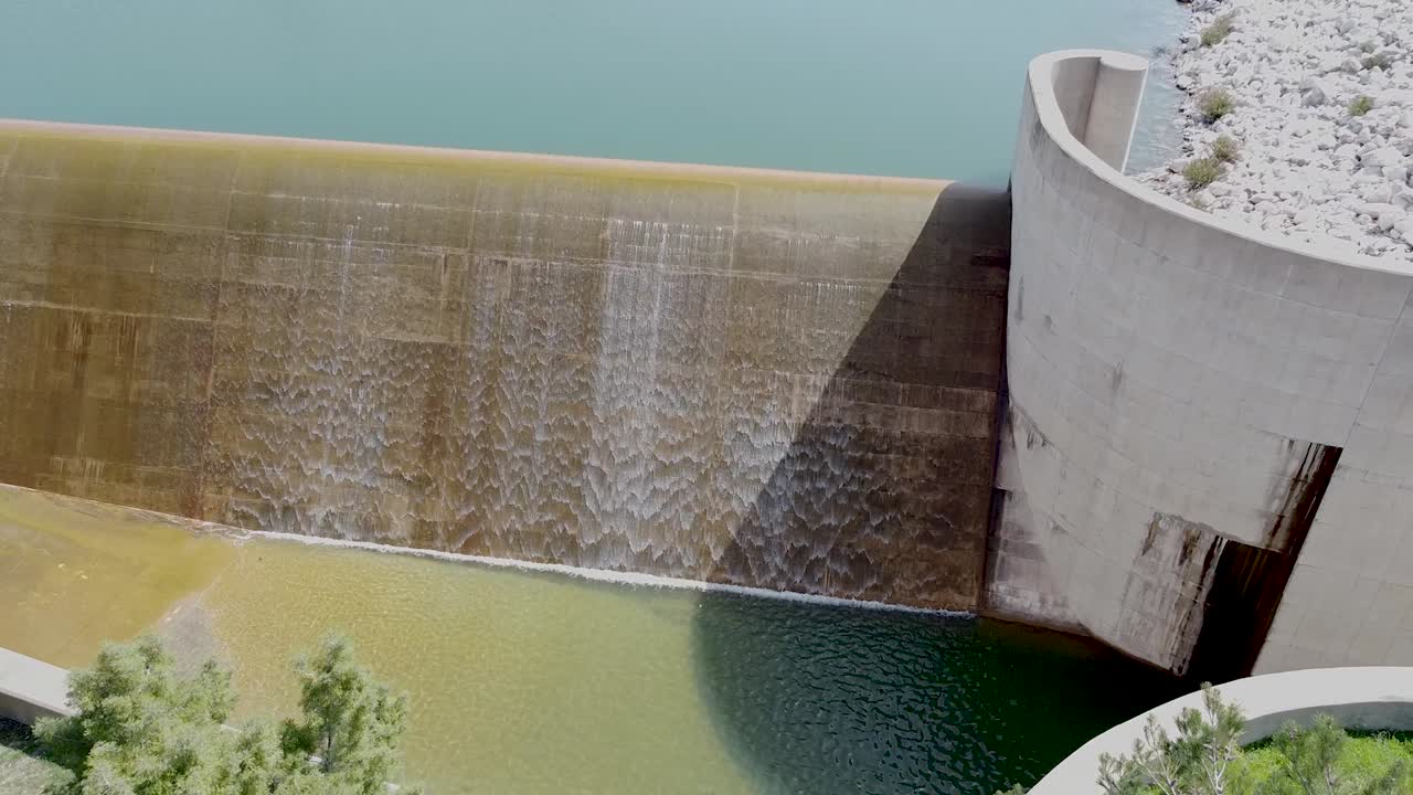 Water rushes over the edge of a large dam, creating a mesmerizing cascade under bright sunlight. The tranquil setting highlights the beauty of nature and engineering