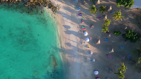 Drone-Desciende-Sobre-Largas-Sombras-De-Palmeras-Y-Sombrillas-En-La-Escondida-Playa-De-La-Cala-Tropical-De-Daaibooi-Curacao