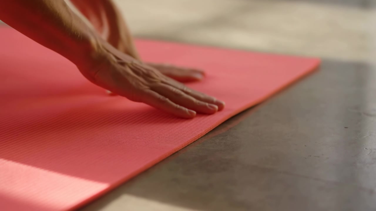 Close-up view of a hand adjusting a red yoga mat on a smooth floor in a calm indoor setting, with soft natural light and a clean wellness atmosphere