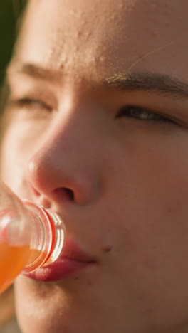 primer plano de una mujer bebiendo jugo de naranja de una botella de plástico al aire libre, reflejando satisfacción, la luz del sol brillando en su cara y la botella, con un suave resplandor de luz y vegetación en el fondo