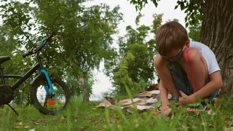 a young child sits under a tree, deep in thought, with a visible bruise on his leg, his hands rest on his legs as he reflects, with a parked blue bicycle nearby and a blanket beneath him