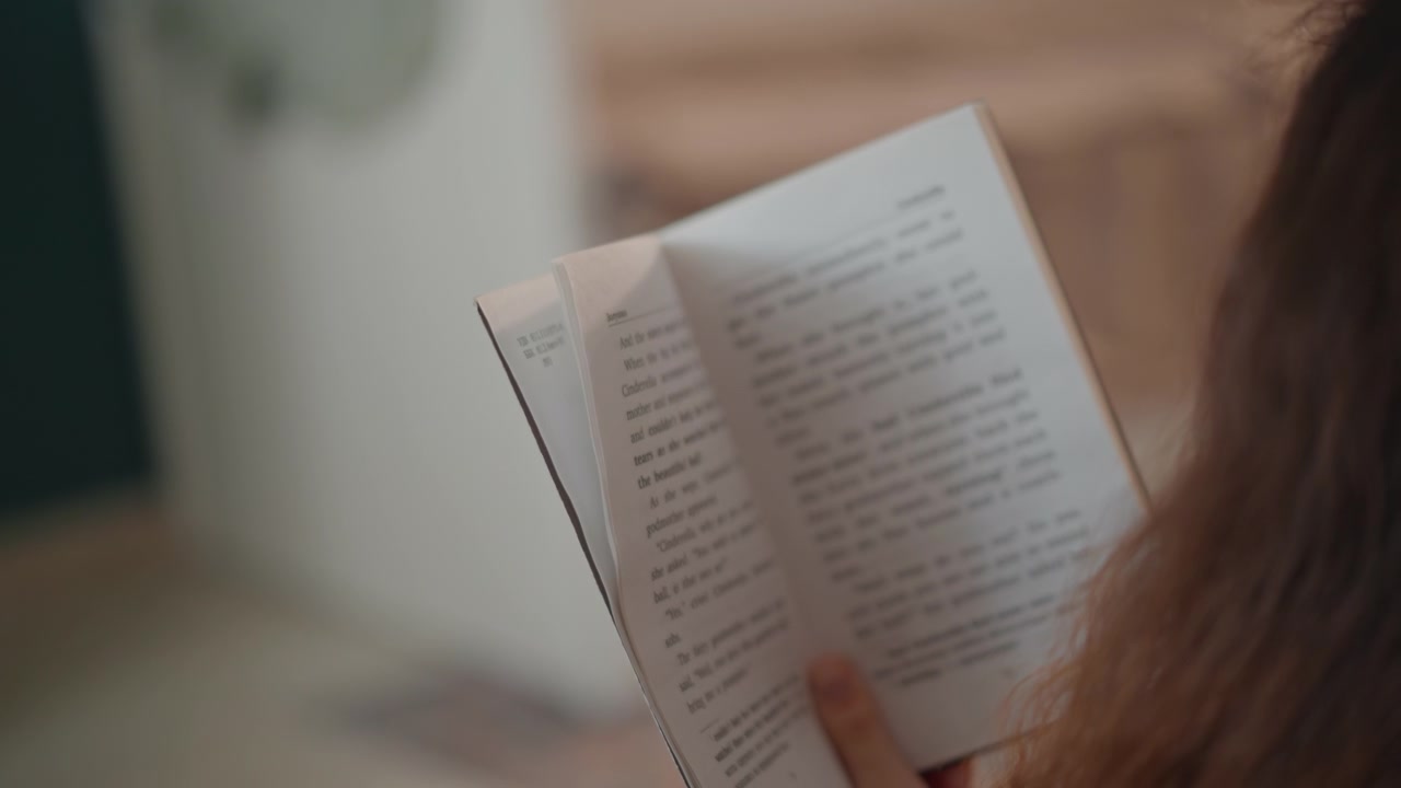 Vista da sopra la spalla di una persona che legge un libro tascabile mentre gira pagina, le punte delle dita sollevano l'angolo con un movimento delicato, luce naturale soffusa, ambiente interno accogliente, atmosfera di vita calma e concentrata.