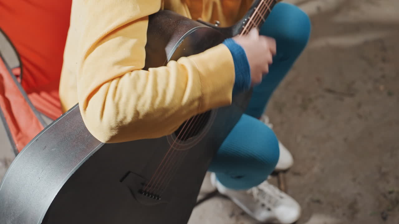 Bredt skud af en campist, der spiller akustisk guitar i en campingomgivelser. En orange campingstol og sandet jord er synlig. Personen har en afslappet holdning med hvide sneakers, et muntert anslag og en jævn akkordprogression til en udendørs jam.