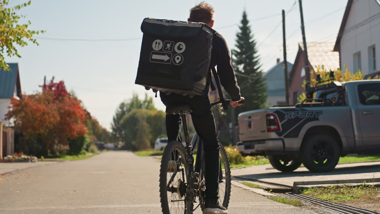 Nærbillede af cyklist, der leverer i nabolaget og cykler forbi en pickup truck og huse, med en fremtrædende brandet isoleret taske, skarpt efterårslys, fokuseret bevægelse og kadence, gig-økonomi-bud, der udfører lastmile