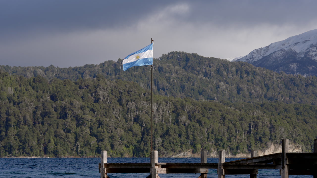 Argentinian flag waving on wooden pier over Patagonian lake with forested mountains backdrop.
